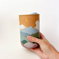 Mountains Travel Mug, a ceramic mug with a matte finish, with a mountain scape around the perimeter. Comes with a blue silicone lid.