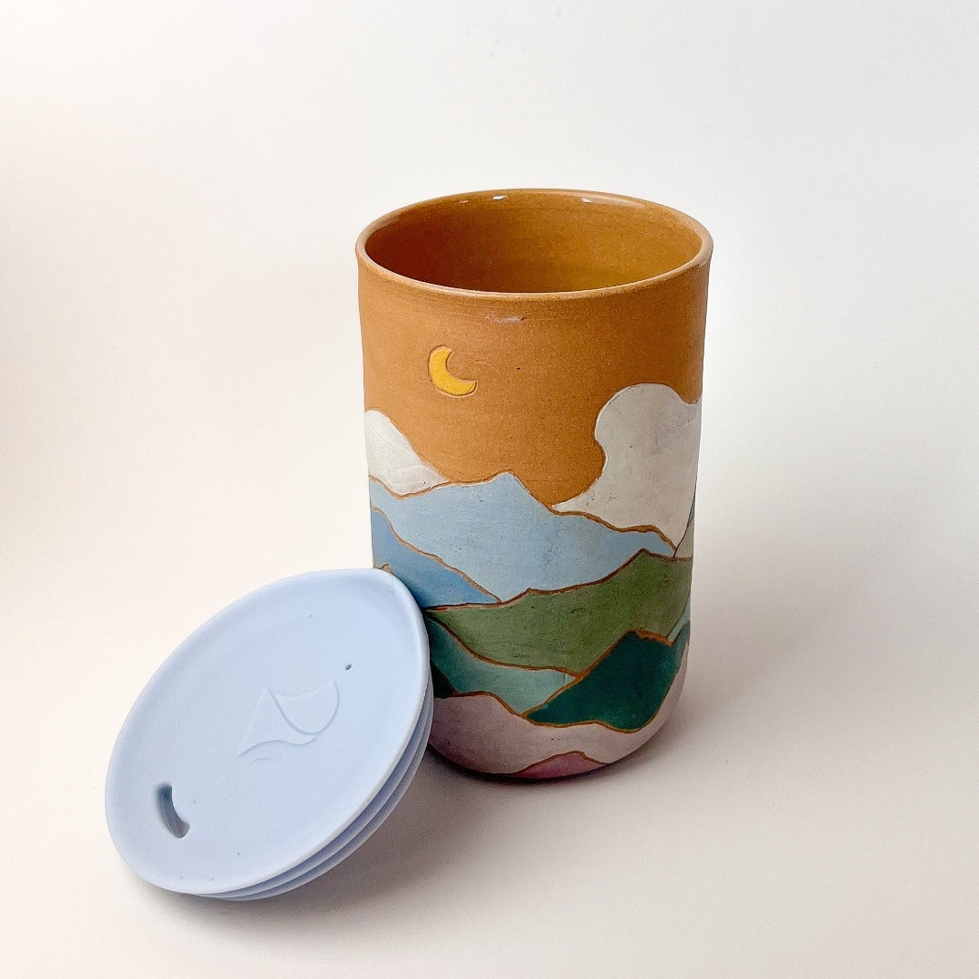 Mountains Travel Mug, a ceramic mug with a matte finish, with a mountain scape around the perimeter. Comes with a blue silicone lid.