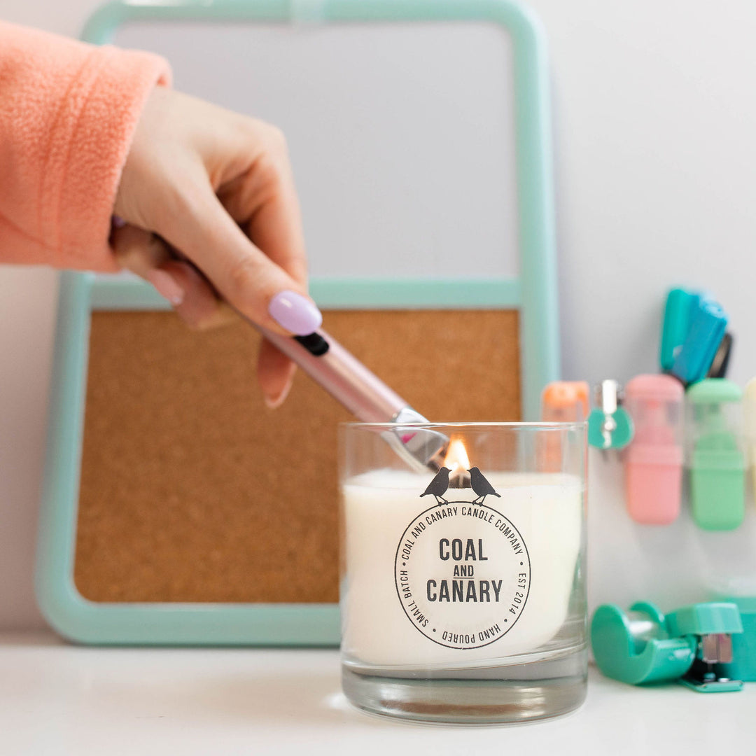 a wood wick candle in a glass jar with the Coal & Canary logo, being lit by an electric lighter.