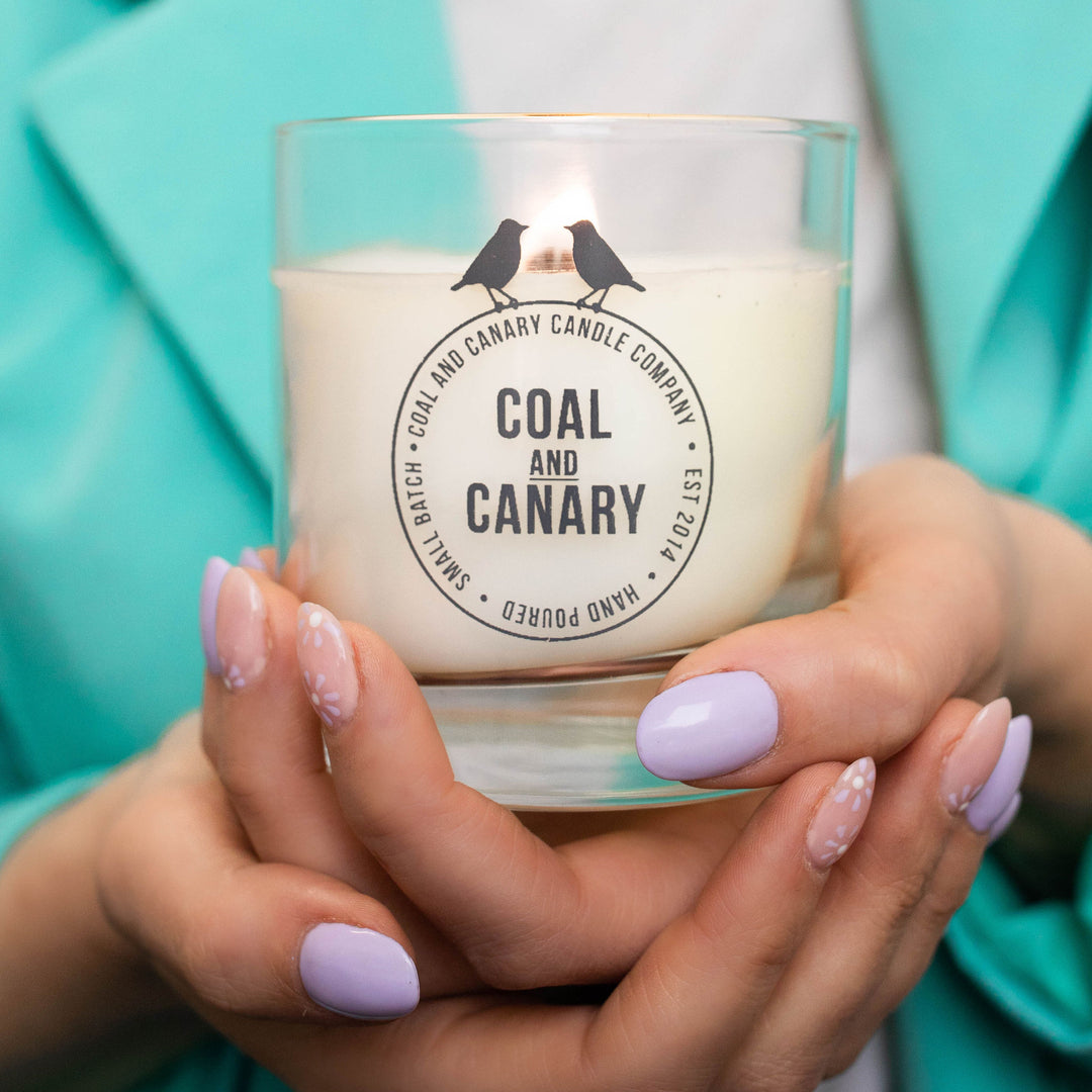 a wood wick candle in a glass jar with the Coal & Canary logo.