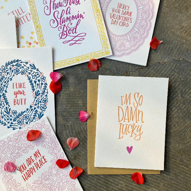 Collection of humorous Valentine's Day cards on a wooden surface with red rose petals.
