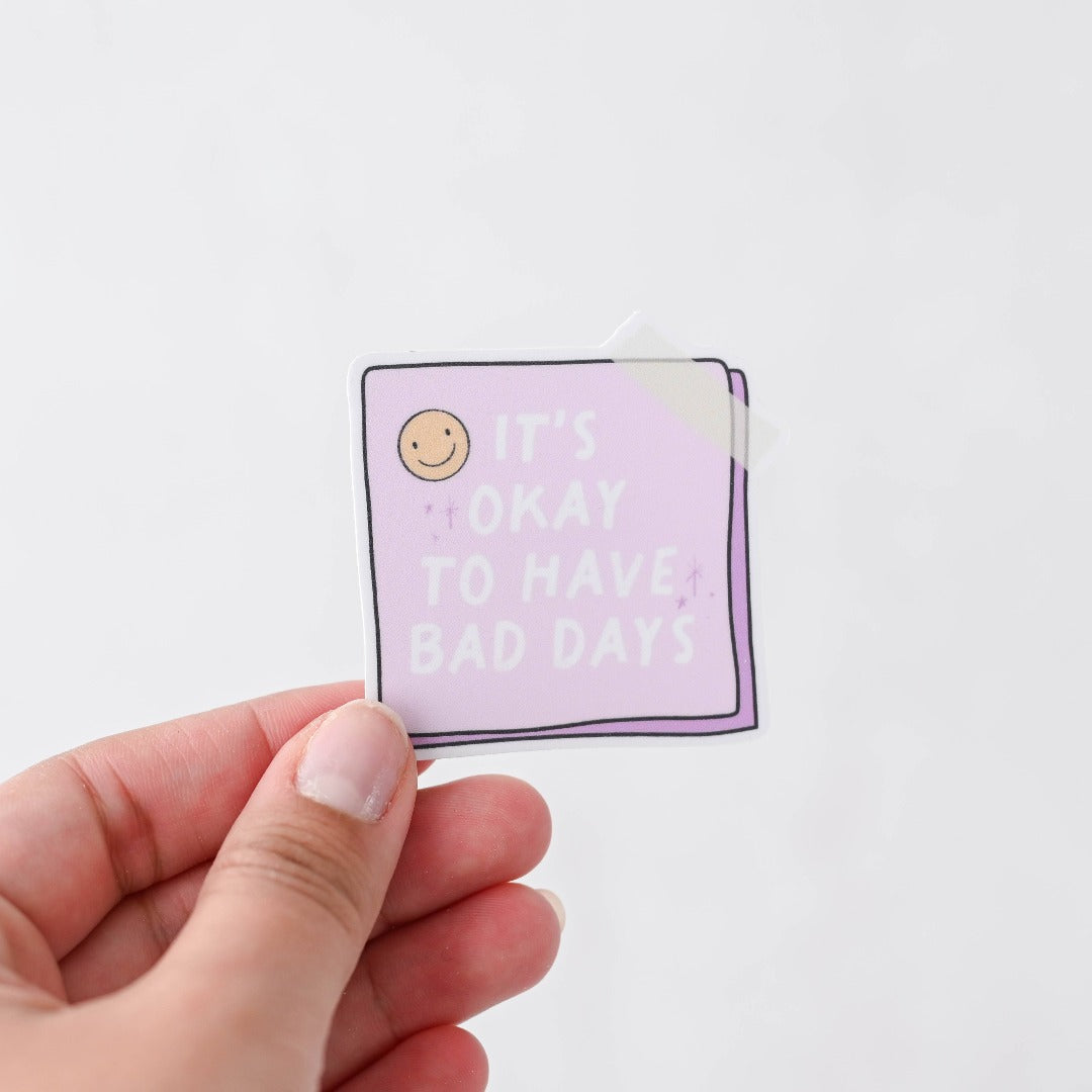 A hand holding a It's Okay To Have Bad Days Sticker, a white sticker with a purple post it reading "It's okay to have bad days"