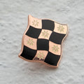 Rose gold-toned pin shaped like a checkered flag with black and white squares. Each square has a small, white flower design in the center.