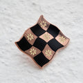 Rose gold-toned pin shaped like a checkered flag with black and white squares. Each square has a small, white flower design in the center.