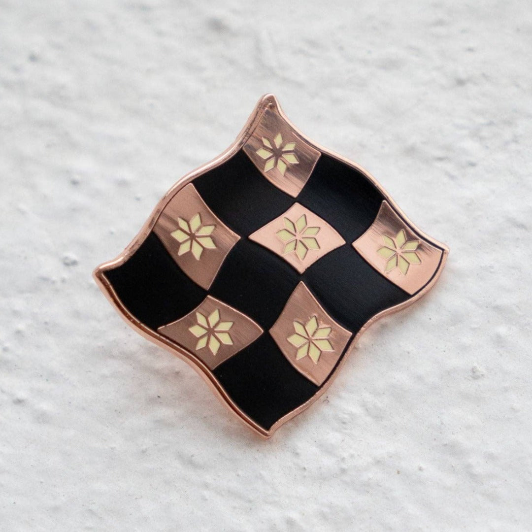 Rose gold-toned pin shaped like a checkered flag with black and white squares. Each square has a small, white flower design in the center.