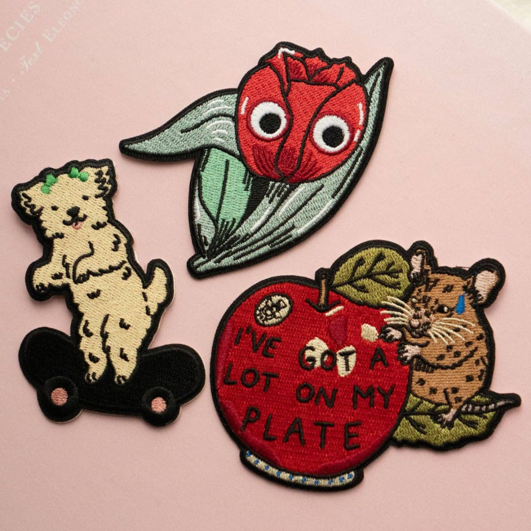 Set of four colorful patches featuring a dog, a pea pod, an apple with text, and a mouse on a pink background.