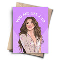 A humorous birthday card with a cartoon drawing of Jennifer Lopez in a glamorous outfit. The text at the top of the card reads, "YOU AGE LIKE J.LO." A brown kraft envelope is behind the card.