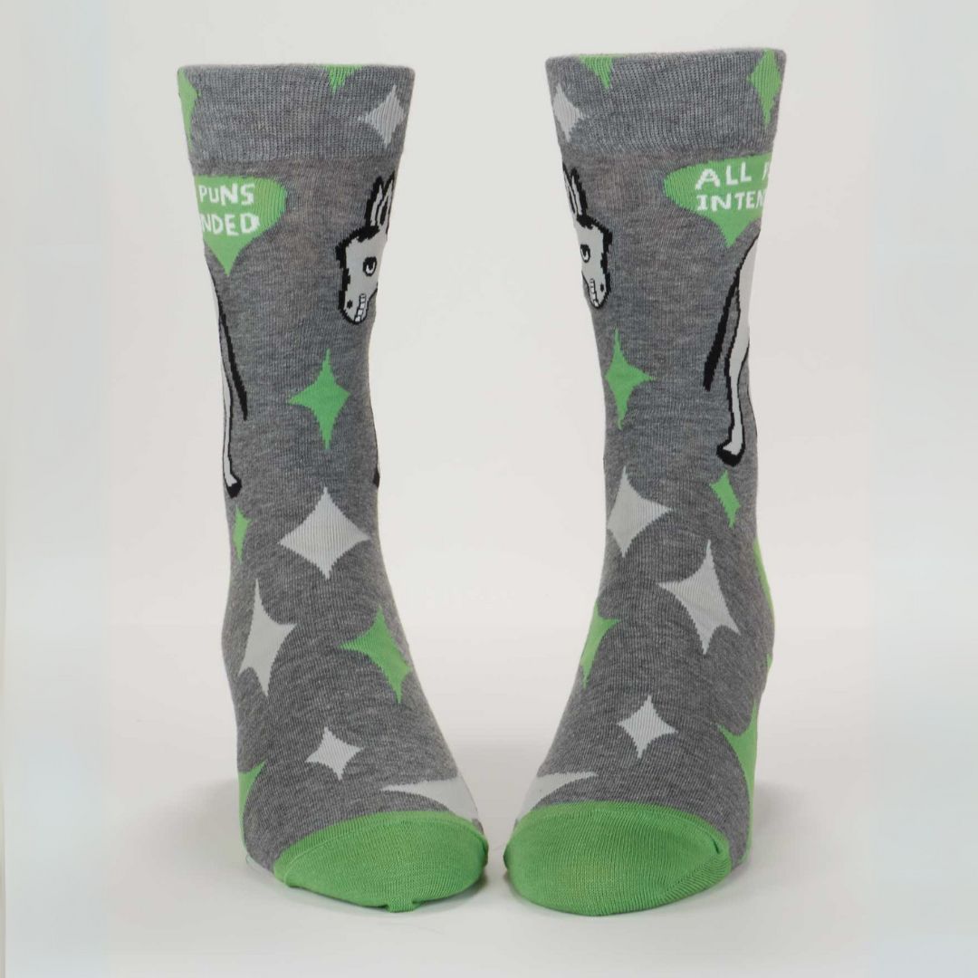A pair of men's crew socks with a grey background and a pattern of white starbursts and green accents. One sock features the rear end of a white donkey and the text "ALL PUNS INTENDED," while the other shows the front half of the donkey.