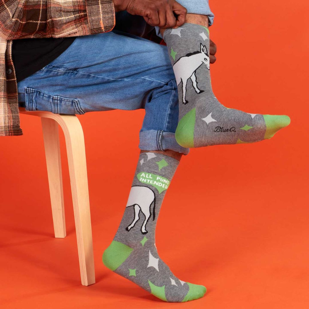 A pair of men's crew socks with a grey background and a pattern of white starbursts and green accents. One sock features the rear end of a white donkey and the text "ALL PUNS INTENDED," while the other shows the front half of the donkey.