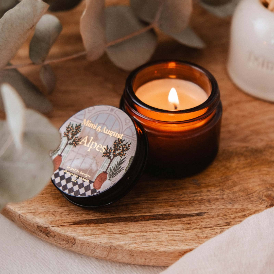 Candle in a glass jar with a decorative label on a wooden surface, surrounded by eucalyptus leaves.