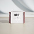 A bar of Aide Bodycare Amber Woodsmoke Soap, featuring a dark purple-brown color and a paper label with the 'aide' logo and 'Amber Woodsmoke' text, displayed on a light-colored surface with a shadow overlay. The soap is natural and handmade, available in Ottawa, Ontario, Canada.