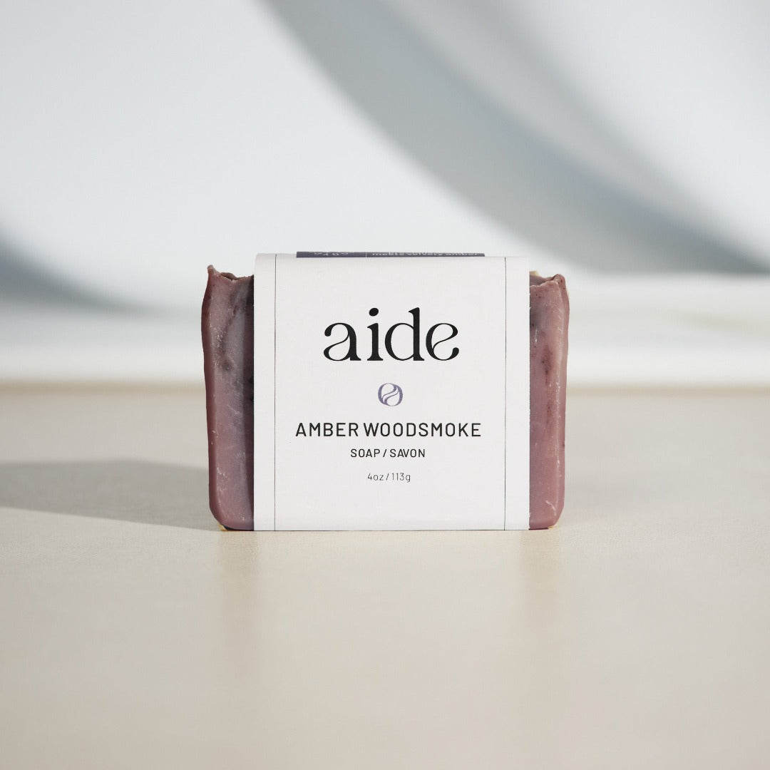 A bar of Aide Bodycare Amber Woodsmoke Soap, featuring a dark purple-brown color and a paper label with the 'aide' logo and 'Amber Woodsmoke' text, displayed on a light-colored surface with a shadow overlay. The soap is natural and handmade, available in Ottawa, Ontario, Canada.