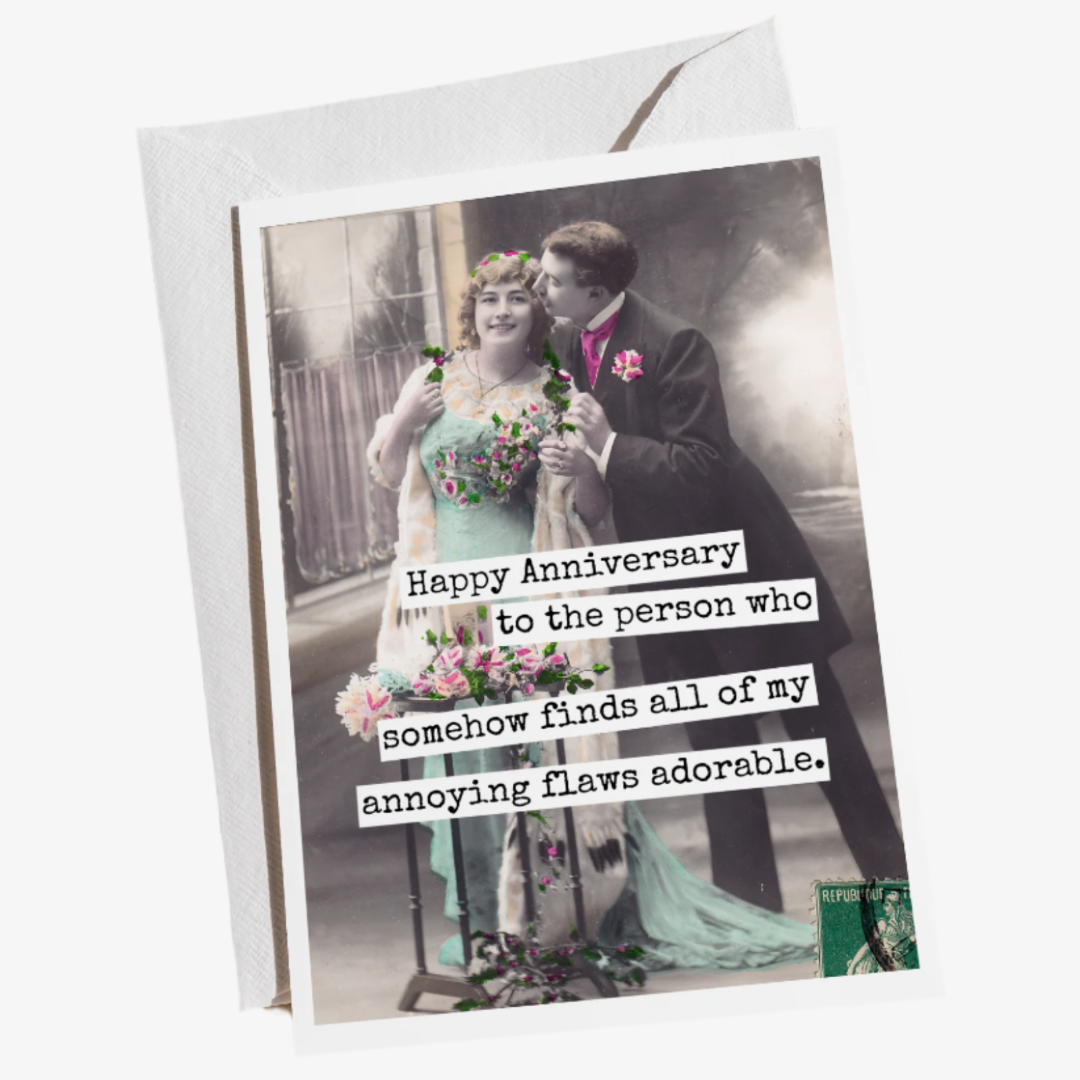 An anniversary card with a vintage photo of a man in a suit embracing a woman in a light blue dress, both holding flowers. Text on the right side of the card reads: "Happy Anniversary to the person who somehow finds all of my annoying flaws adorable." The card is resting on a textured cream-coloured fabric with some wispy dried grasses.