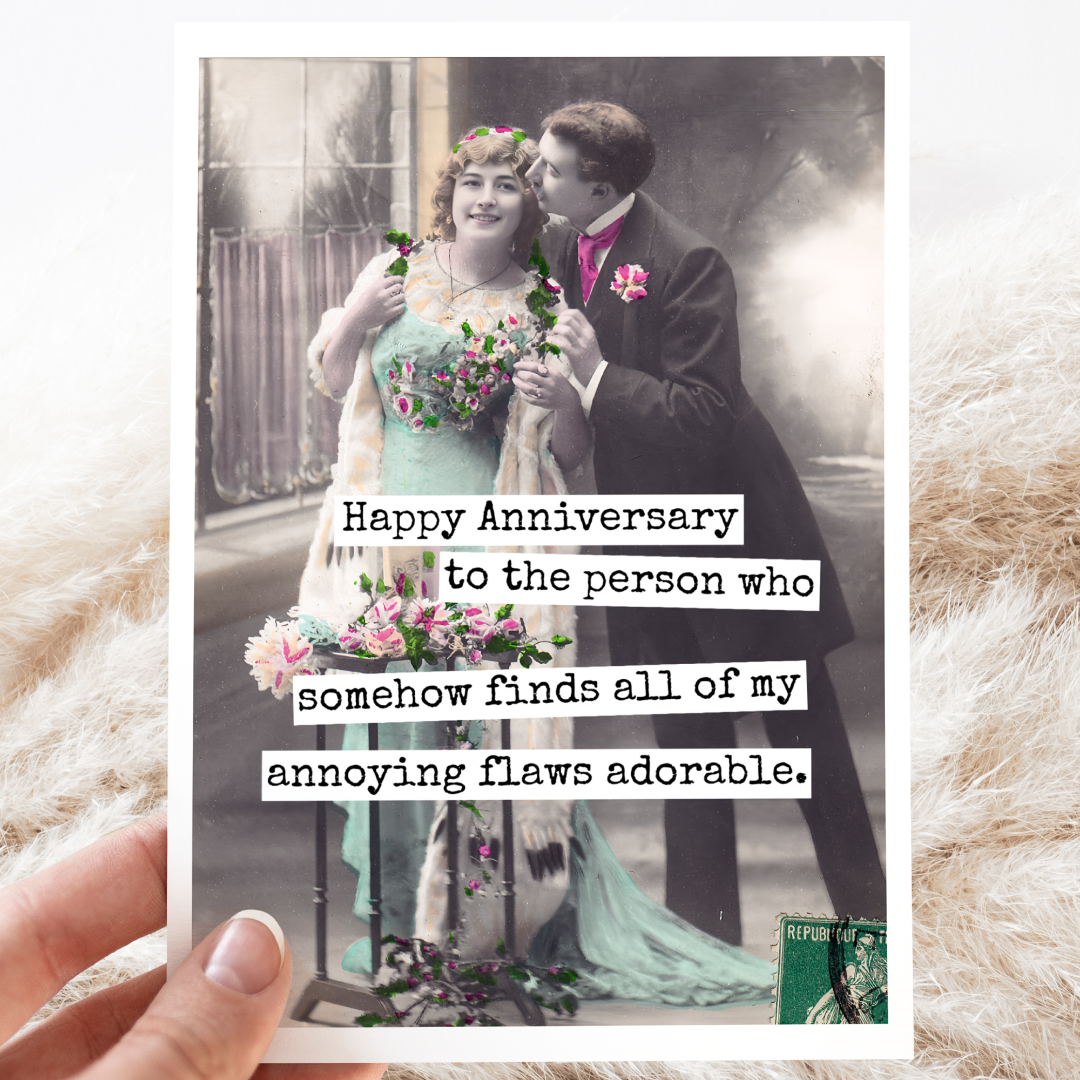 An anniversary card with a vintage photo of a man in a suit embracing a woman in a light blue dress, both holding flowers. Text on the right side of the card reads: "Happy Anniversary to the person who somehow finds all of my annoying flaws adorable." The card is resting on a textured cream-coloured fabric with some wispy dried grasses.