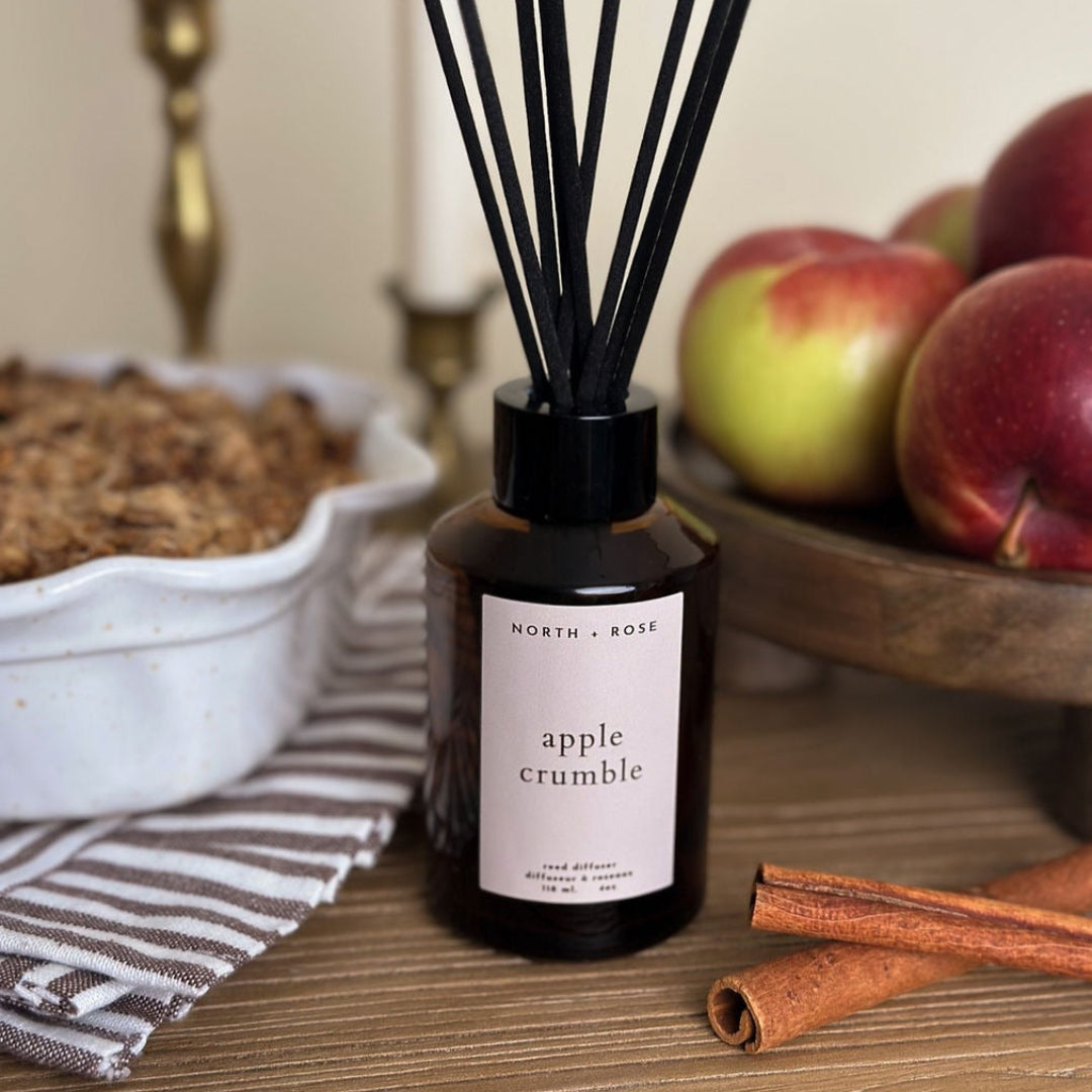 Diffuser bottle labeled 'apple crumble' with apples and cinnamon sticks on a table.