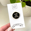 Enamel pin with 'i make bad choices' text on a card, held by a hand with a plant in the background.