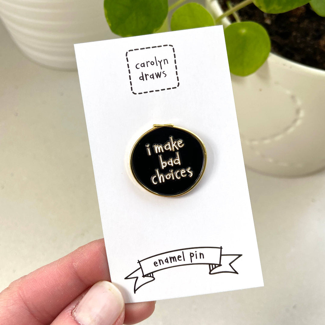 Enamel pin with 'i make bad choices' text on a card, held by a hand with a plant in the background.