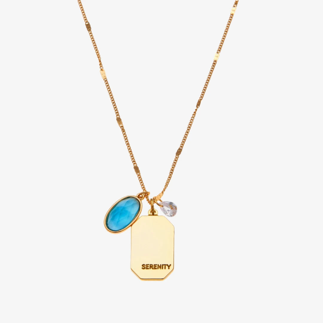 A gold necklace with an aquamarine stone pendant. The pendant also has the word "SERENITY" engraved on it. The necklace is displayed on a light blue card with the month "March" and the brand name "Foxy Originals."