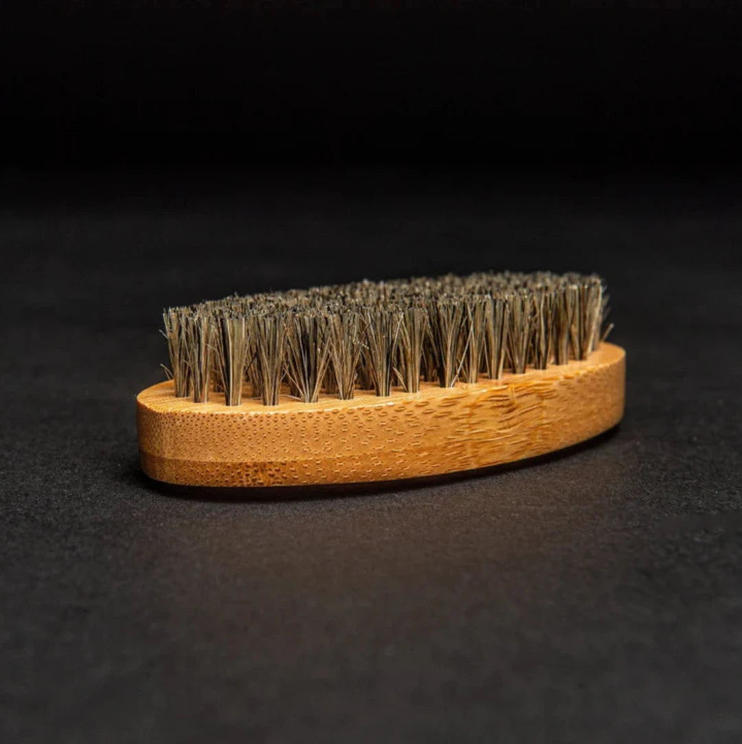 A brown, oval-shaped brush with a wooden handle and natural bristles. The brush is resting on a black surface.