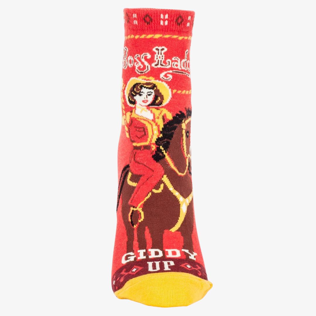 A pair of red ankle socks with yellow accents. One sock features a vintage-style illustration of a cowgirl riding a horse, with "Boss Lady" text above and "Giddy Up" below. The other sock shows the back of the cowgirl and horse with the "Giddy Up" text.