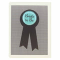 A wedding card with a black and white striped background. A black ribbon shape is centered on the card, and on top of it is a round, light blue button with the words "Bride to Be" in dark blue script.