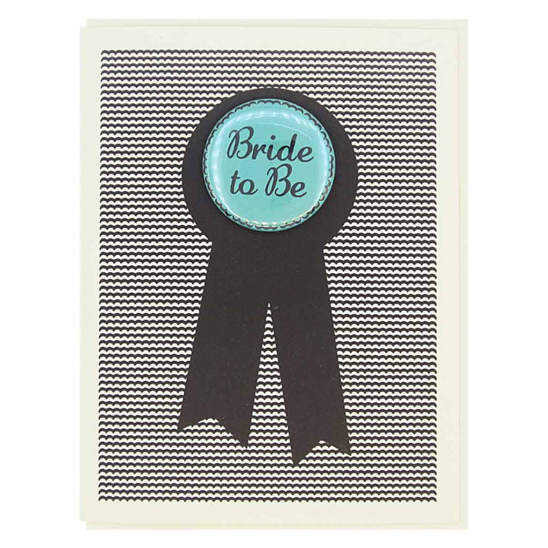 A wedding card with a black and white striped background. A black ribbon shape is centered on the card, and on top of it is a round, light blue button with the words "Bride to Be" in dark blue script.