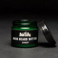 A dark green glass jar with a black lid. The jar is filled with beard butter and has a black label with the text "BUSH BEARD BUTTER SWEET" on it. The lid is slightly open and the jar is resting on a black surface.