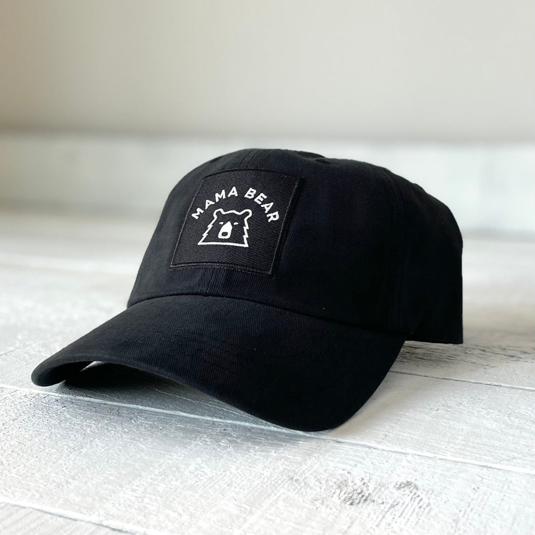 A black baseball cap with a low profile and curved peak, featuring a white square patch with a black bear face and the text "MAMA BEAR" in white. Shop this at Cheerfully Made, Canadian-made gift shop.