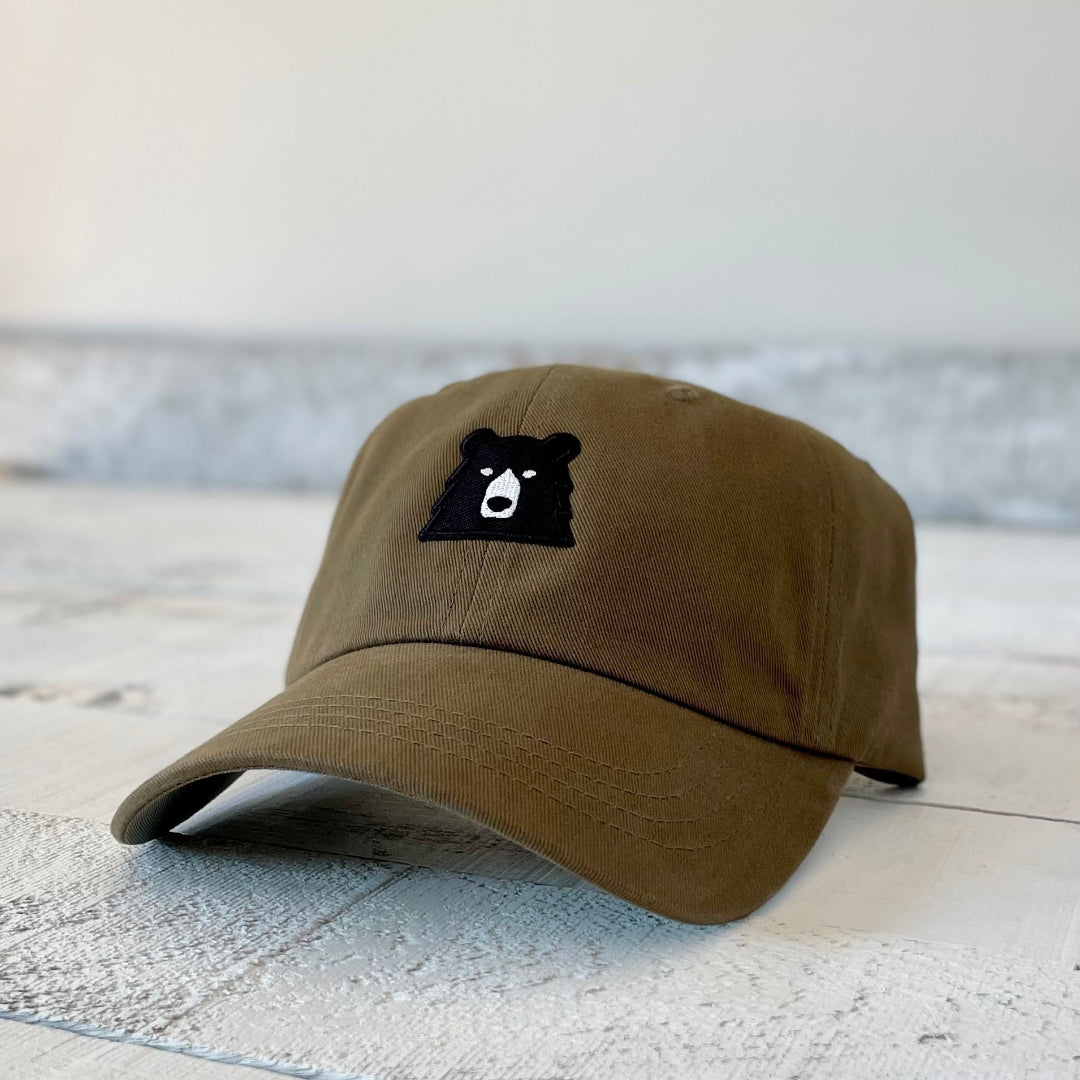 A peat (dark brown/green) baseball cap with a low profile and curved peak, featuring a cute black bear face patch with white eyes and snout stitched on the front. Shop this at Cheerfully Made, Canadian-made gift shop.
