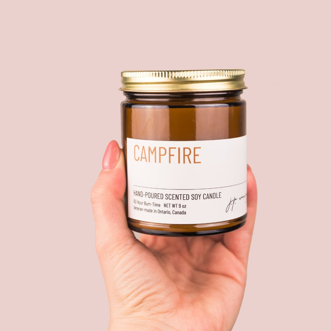 Hand holding a 'Campfire' scented soy candle with a gold lid against a beige background