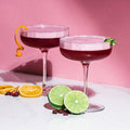 Two cocktails with garnishes on a white surface with a pink background
