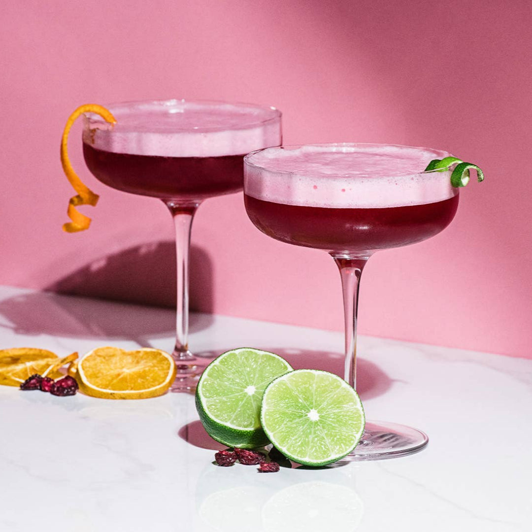 Two cocktails with garnishes on a white surface with a pink background