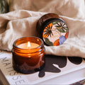 Two amber glass candles with decorative lids on a light fabric background