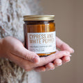 Hand holding a candle labeled 'Cypress and White Pepper' against a neutral background