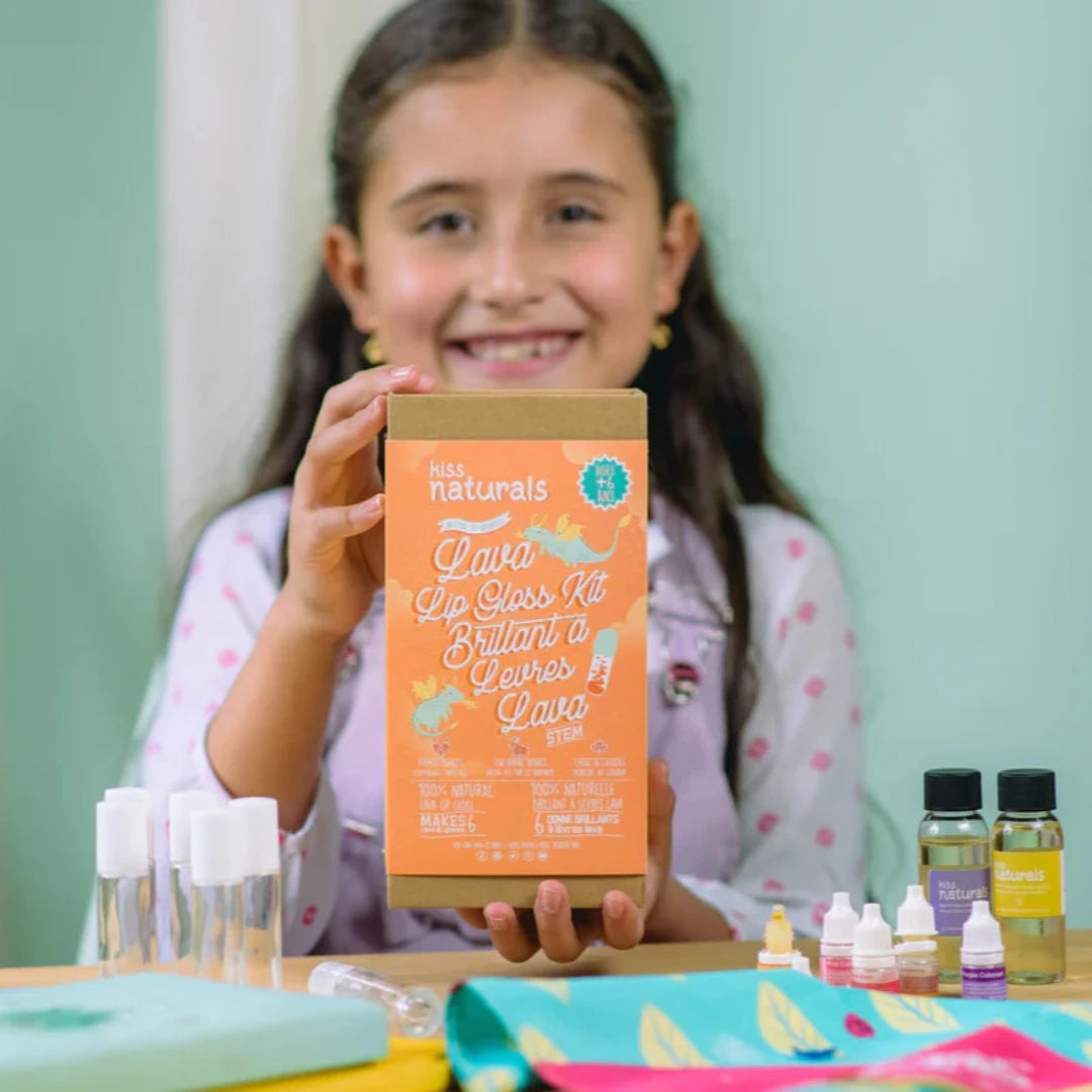 A cardboard box containing a Kiss Naturals Lava Lip Gloss Kit. The box is orange and white with the product name and a colorful illustration of a dragon. It states that the kit is 100% natural, made in Canada, and suitable for ages 6 and up.