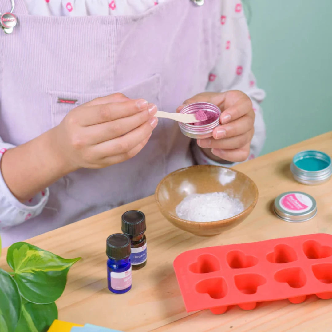 A cardboard box containing a Kiss Naturals Bath Bomb Kit. The box is green and white with the product name and a colorful illustration of bath bombs. It states that the kit is 100% natural, made in Canada, and suitable for ages 6 and up.
