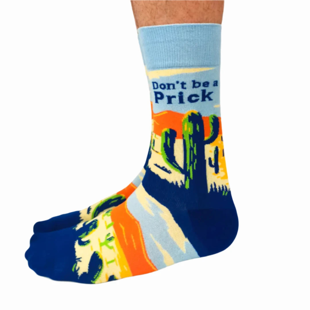 A pair of crew socks with a desert scene and the text "Don't Be a Prick." The socks are mostly light blue with a darker blue heel and toe area. The desert scene is depicted in orange, yellow, and green.