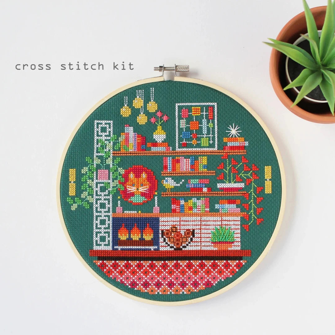 A completed cross-stitch project in a wooden hoop. The design depicts a cozy living room scene with a fireplace, bookshelves, plants, and a cat. The scene is filled with colorful details and is set against a green background. The text "cross stitch kit" is written above the hoop