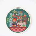 A completed cross-stitch project in a wooden hoop. The design depicts a cozy living room scene with a fireplace, bookshelves, plants, and a cat. The scene is filled with colorful details and is set against a green background. The text "cross stitch kit" is written above the hoop