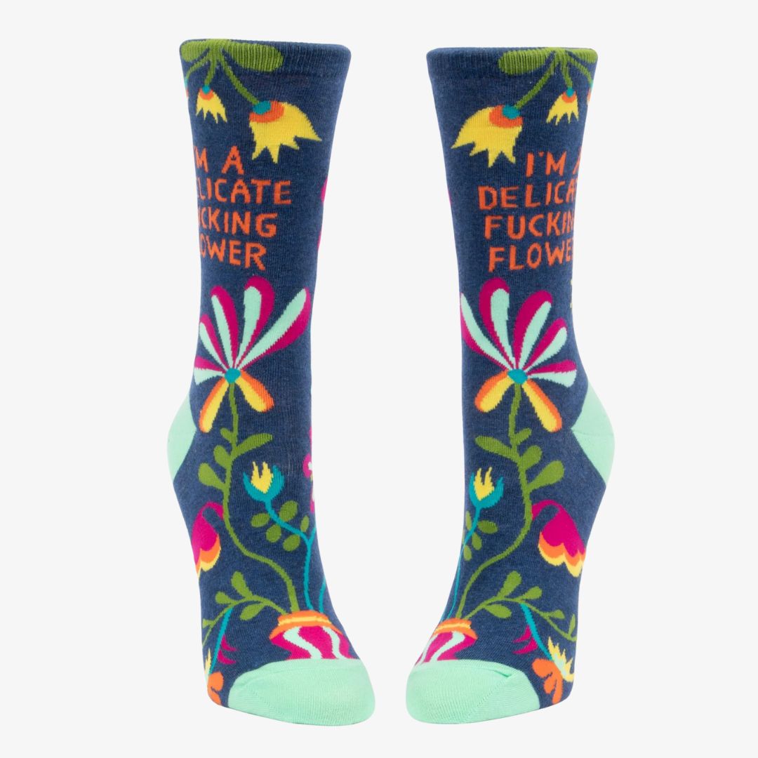 A pair of dark blue women's crew socks with a vibrant, abstract floral pattern in shades of orange, yellow, pink, fuchsia, green, and light blue. Text on one sock reads "I'M A DELICATE FUCKING FLOWER".