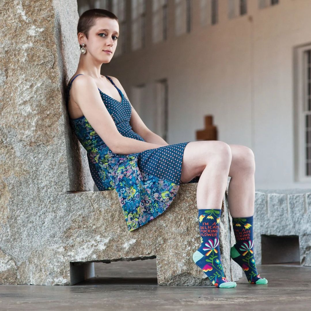 A pair of dark blue women's crew socks with a vibrant, abstract floral pattern in shades of orange, yellow, pink, fuchsia, green, and light blue. Text on one sock reads "I'M A DELICATE FUCKING FLOWER".