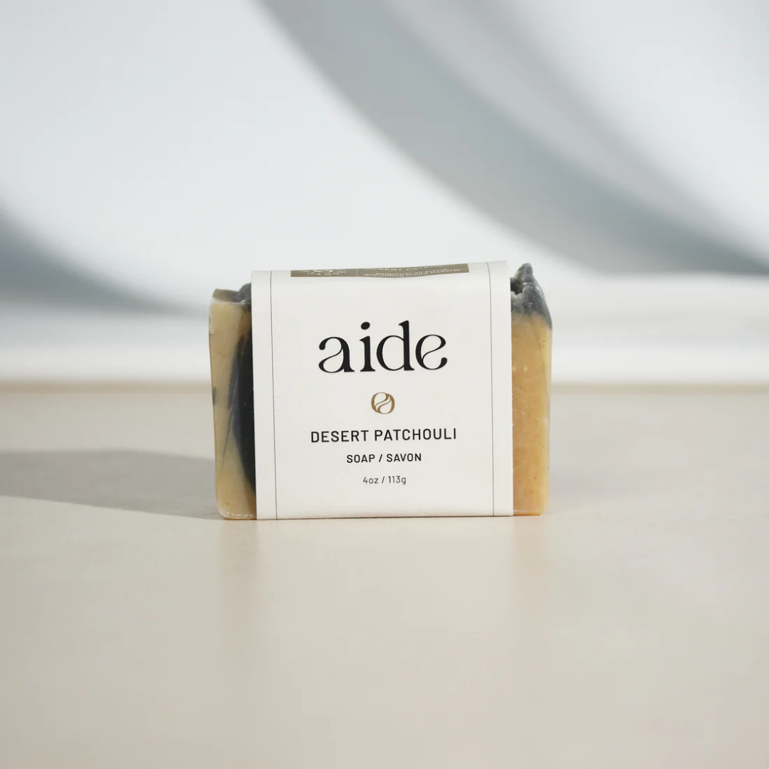 A bar of Aide Bodycare Desert Patchouli Soap, featuring a light beige and dark gray swirled pattern and a paper label with the 'aide' logo and 'Desert Patchouli' text, displayed on a light-colored surface with a shadow overlay. The soap is natural and handmade, available in Mississippi Mills, Ontario, Canada.