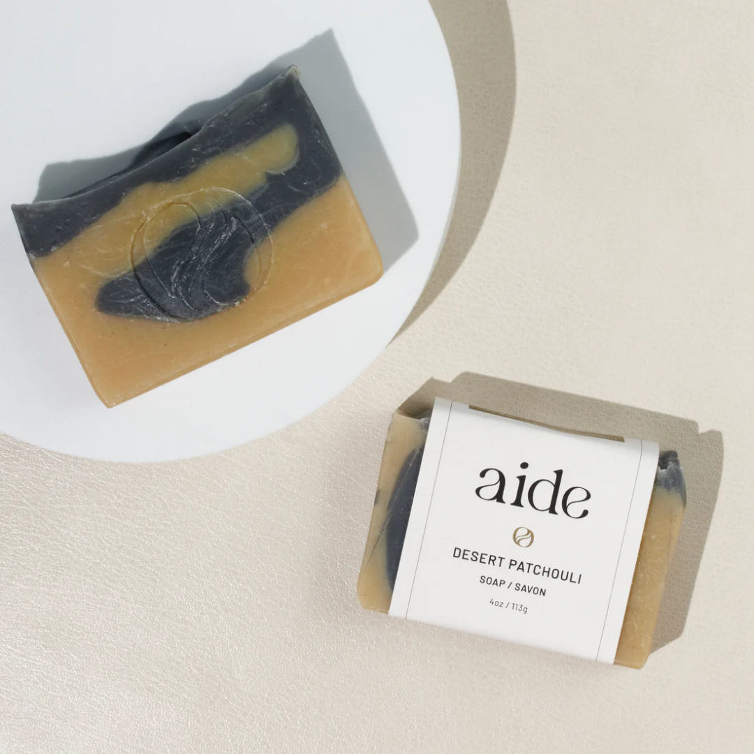A bar of Aide Bodycare Desert Patchouli Soap, featuring a light beige and dark gray swirled pattern and a paper label with the 'aide' logo and 'Desert Patchouli' text, displayed on a light-colored surface with a shadow overlay. The soap is natural and handmade, available in Mississippi Mills, Ontario, Canada.
