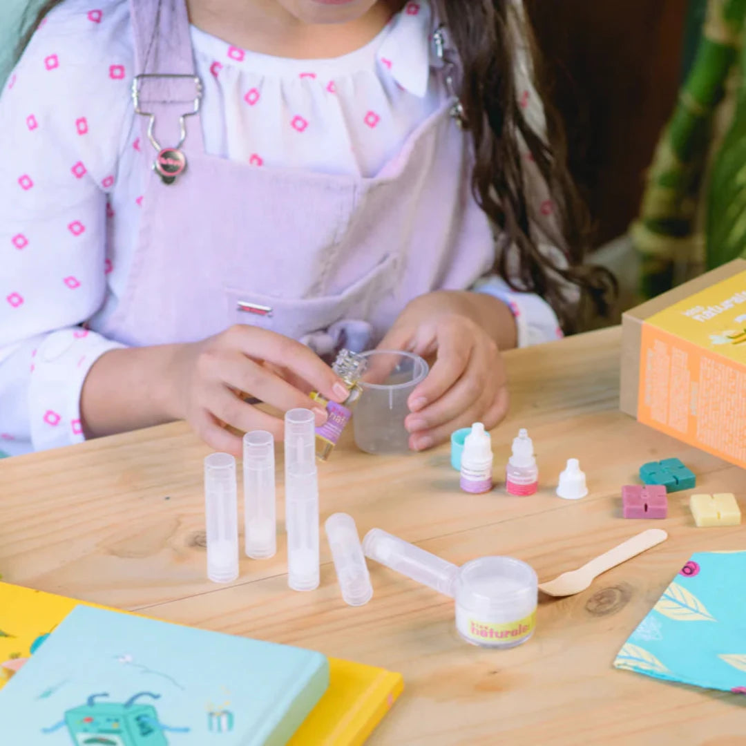 A cardboard box containing a Kiss Naturals Lip Balm Kit. The box is yellow and white with the product name and a colorful illustration of bees. It states that the kit is 100% natural, made in Canada, and suitable for ages 6 and up.