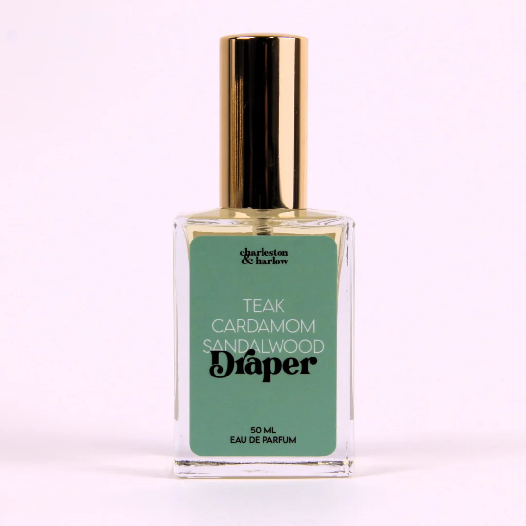A small glass bottle of perfume with a gold cap. The bottle is transparent and filled with a light-colored liquid. The label on the bottle reads "Draper" in large, black letters. Below the brand name, it lists the fragrance notes: "Teak, Cardamom, Sandalwood."