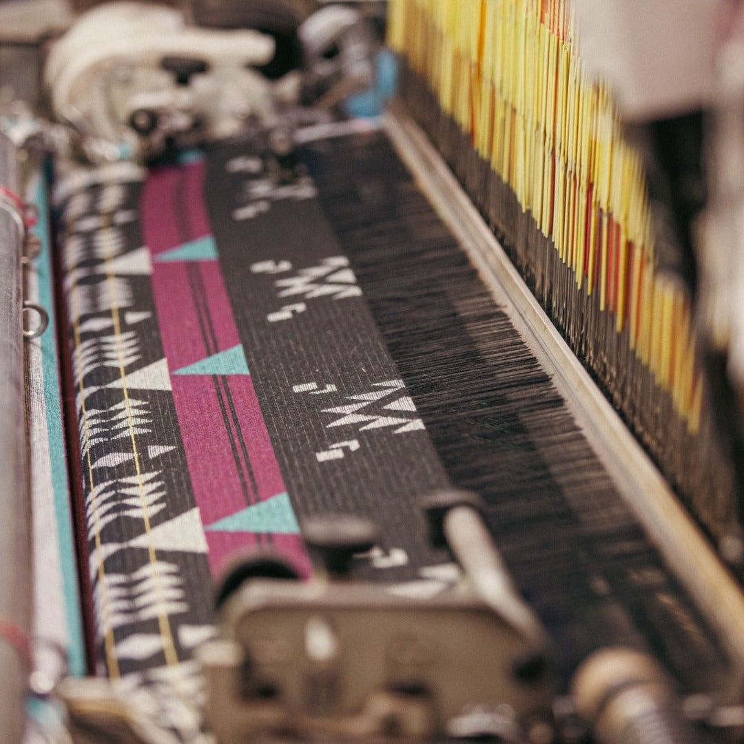 The Éclair Eco-Friendly Everyday Blanket by Mini Tipi, featuring bold Indigenous-inspired geometric patterns in black, grey, burgundy, turquoise, and yellow, on a loom. This sustainable and stylish blanket is available in Ottawa, Ontario, Canada.