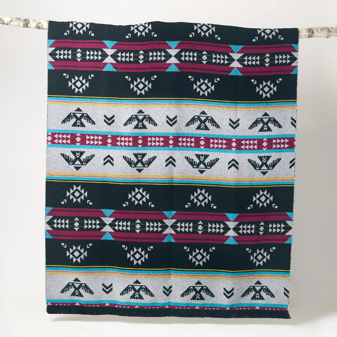 The Éclair Eco-Friendly Everyday Blanket by Mini Tipi, featuring bold Indigenous-inspired geometric patterns in black, grey, burgundy, turquoise, and yellow, hanging from a natural branch. This sustainable and stylish blanket is available in Ottawa, Ontario, Canada.