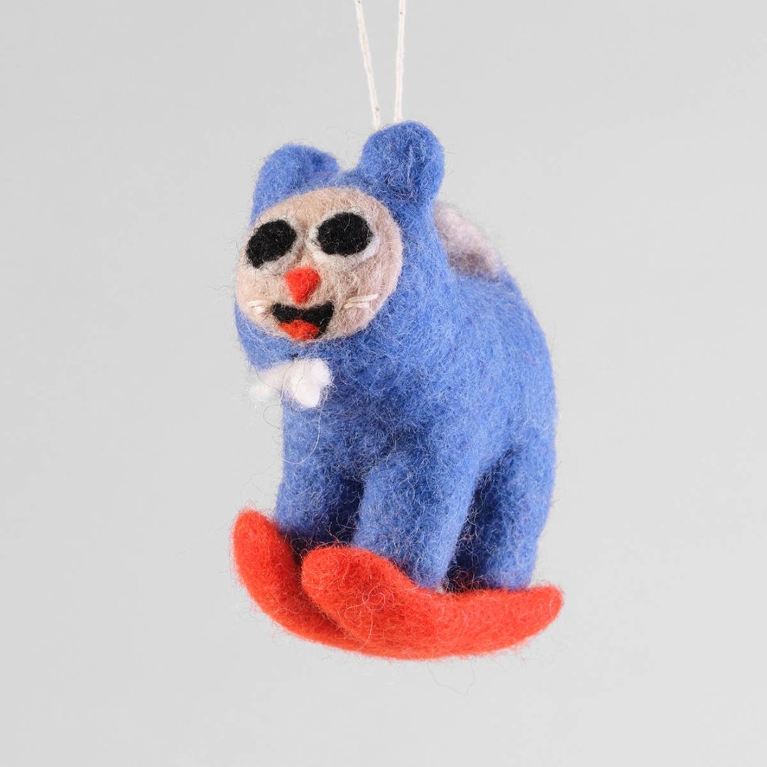 A felt ornament of a cat in a blue snow suit skiing down a hill on red skies. The cat has a white face.