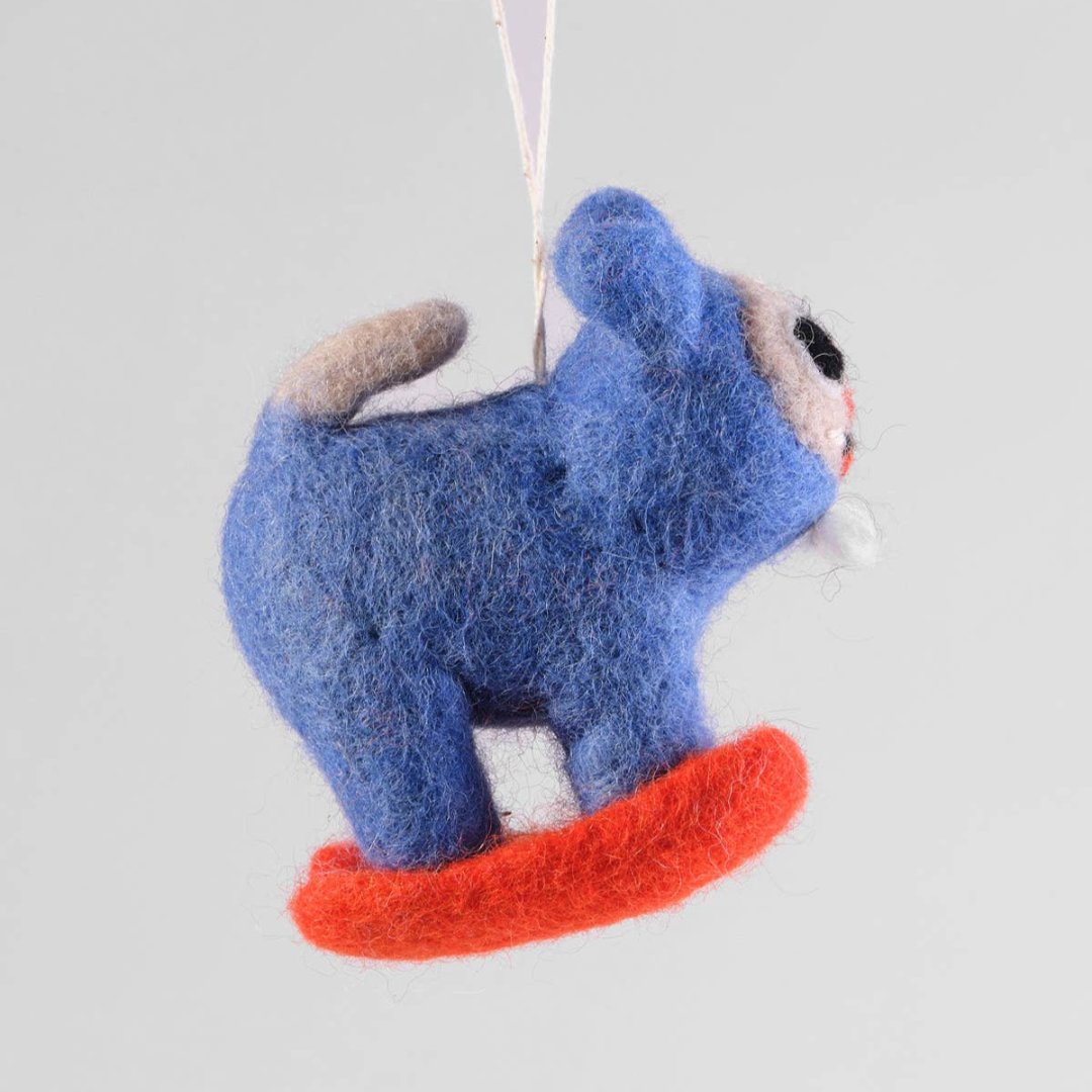 A felt ornament of a cat in a blue snow suit skiing down a hill on red skies. The cat has a white face.