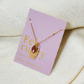 A gold necklace with an amethyst stone pendant. The pendant also has the word "WISDOM" engraved on it. The necklace is displayed on a purple card with the month "February" and the brand name "Foxy Originals."